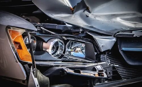 Close-up of the front-end damage to a silver car following a collision, showing a heavily crumpled hood and broken headlight assembly.