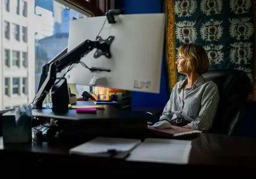 Woman sitting at a office desk with a large computer monitor, looking thoughtfully out a high-rise city window.