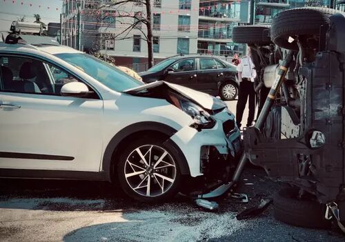 White Kia SUV with heavy front-end damage collided with an overturned vehicle resting on its side at a city intersection.